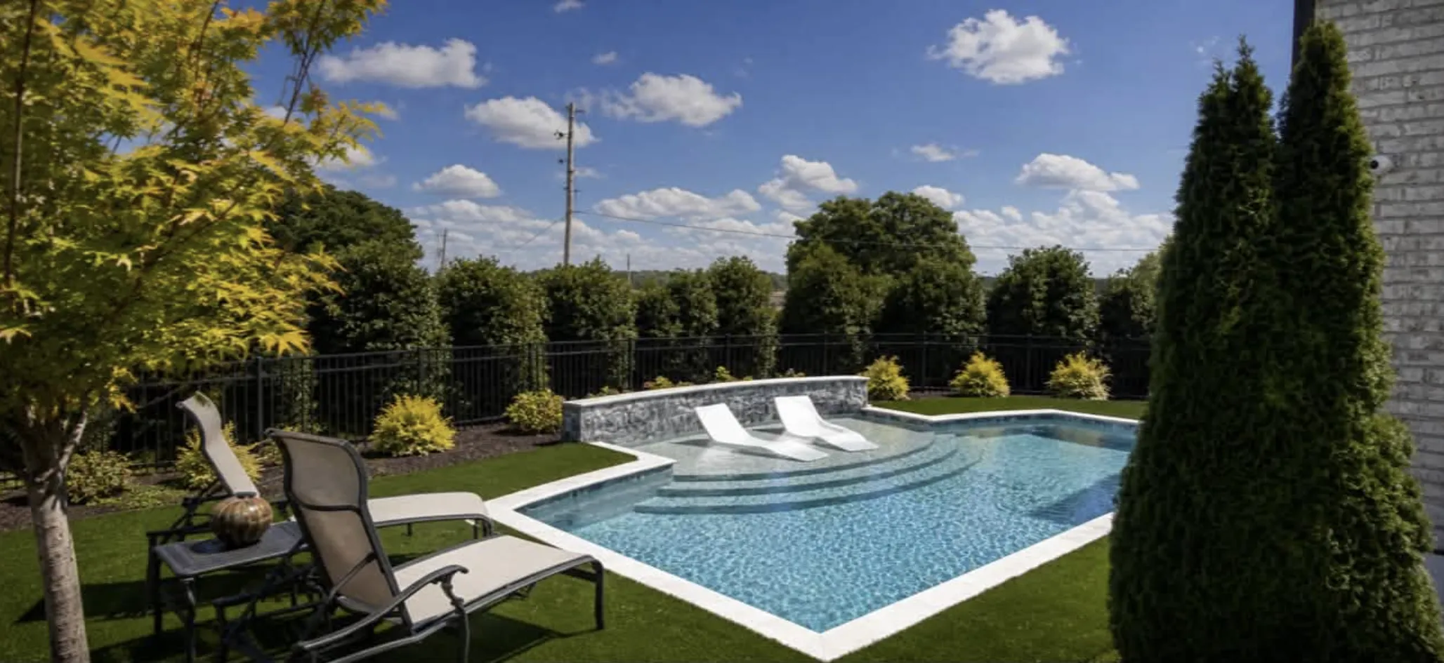 Pool with a built-in tanning ledge and loungers bordered by artificial turf and iron fencing