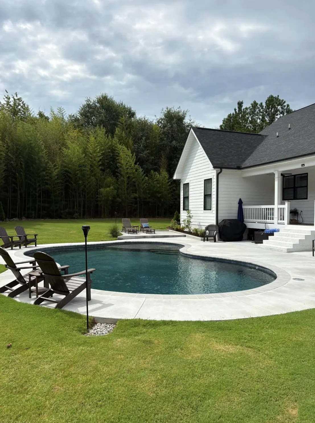 Freeform kidney-shaped pool with a broad concrete deck beside a white farmhouse