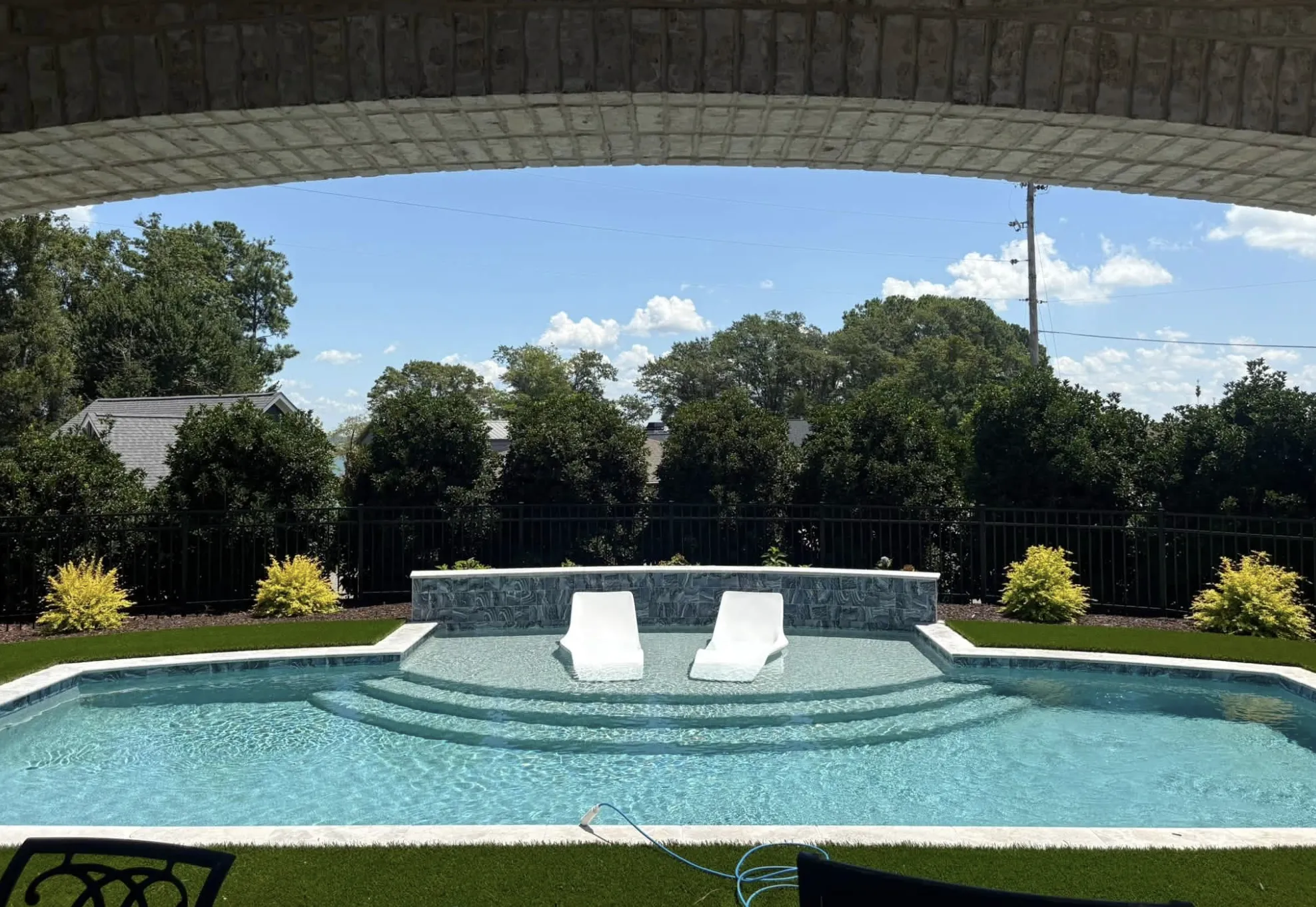 Pool with in-water lounge chairs and a raised stacked-stone wall, framed by a covered stone patio, designed and built by Fireside Pools